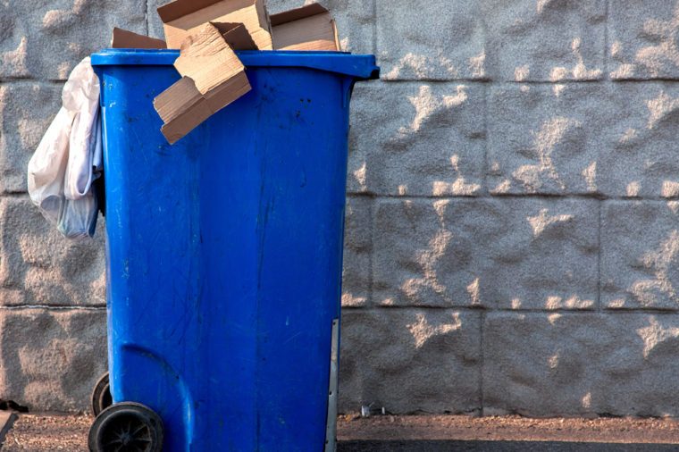 The trash can of blue color filled with a garbage at the top costs on the street near a gray stone wall.