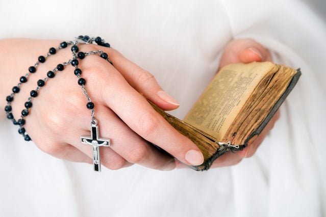 Young woman's hands with a rosary, bible and a white clothing in a background