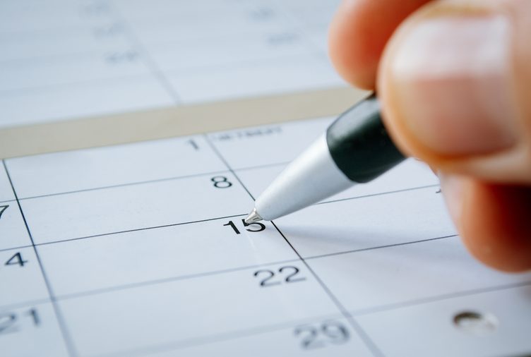 Person writing with a ballpoint pen on a calendar on the date of the 15th with the nib hovering over blank copyspace, oblique angle