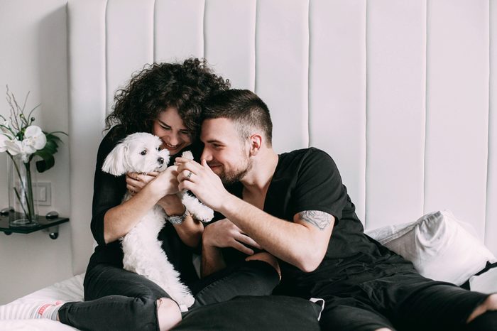 Man and woman in black play with little white dog on bed