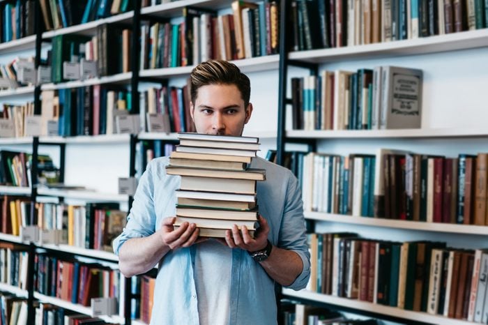 Overworked persistent young man in casual wear holding many literature books in hands for science course work.Intelligent student with textbooks standing in library interior with book shelves