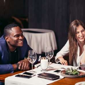 Two interracial young people looking aside at somebody and smiling while siting at the table set for dinner in restaurant