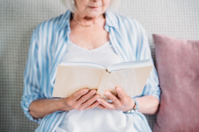 cropped shot of senior woman reading book while resting on sofa at home