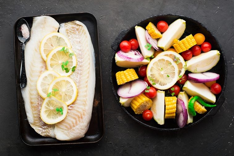 Two dishes before baking: cod with lemons and assorted organic garden vegetables in round form: corn, zucchini, onion, tomatoes, herbs. Top View