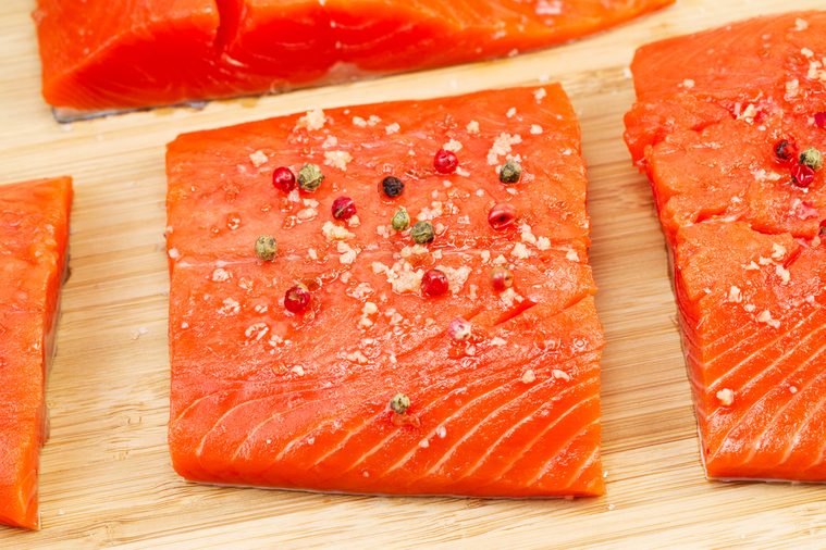 Closeup horizontal photo of dried red peppercorns and sea salt on top of fresh Wild Red Salmon pieces with natural bamboo cutting board underneath 