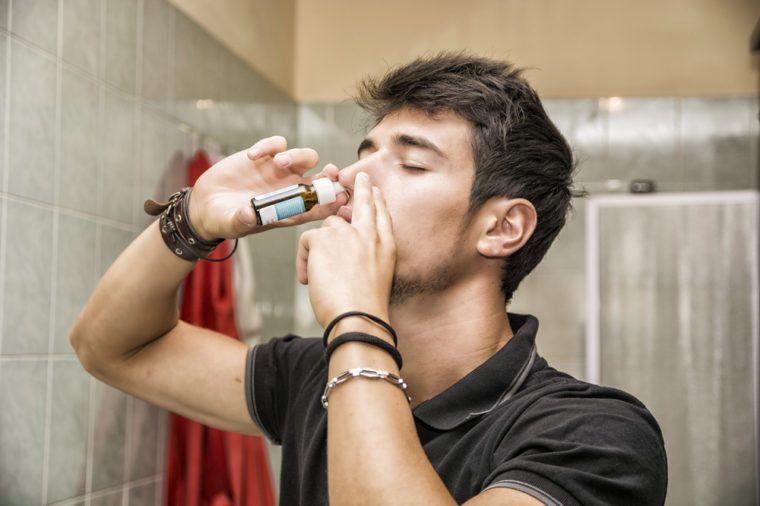 Head and Shoulders Close Up of Young Attractive Man with Dark Hair Sniffing Nose Spray with Eyes Closed in Home Bathroom