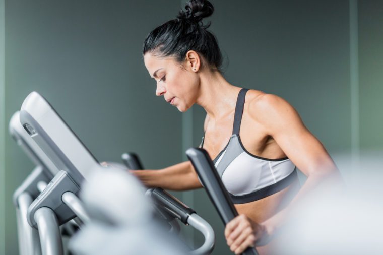 Side view portrait of sweaty fit woman exercising using elliptical machine during intense workout in modern gym