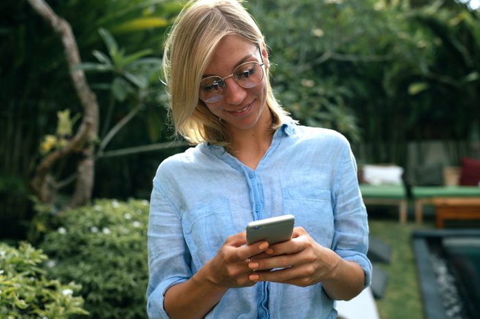 Model look female wearing glasses is reading emails on her mobile phone while relaxing on a luxury tropical villa. Travel blogger wearing light blue shirt is surfing the web on a smartphone.