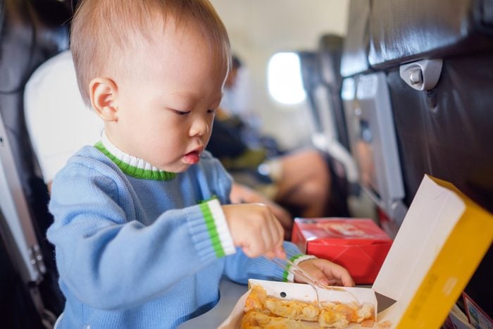 Cute little Asian 18 months / 1 year old toddler baby boy child wearing blue sweater eating food during flight on airplane. Flying with children, Happy air travel with kids & little traveler concept