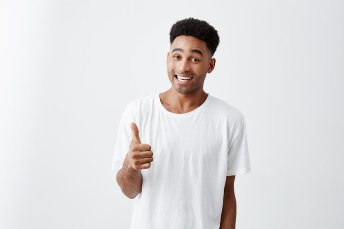 You're looking good, man. Close up portrait of mature good-looking cheerful man with afro hairdo in casual white t shirt smiling with teeth, showing thumb up sign with happy expression.