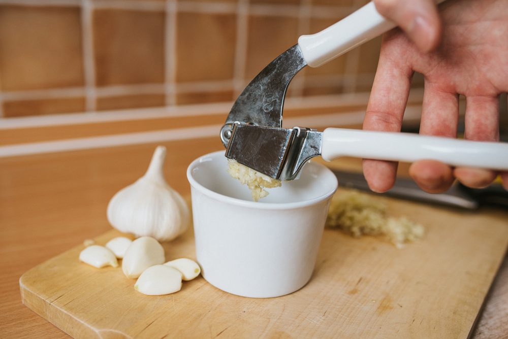 Garlic crushed squeezed from garlic press with white bowl and wooden cutting board.