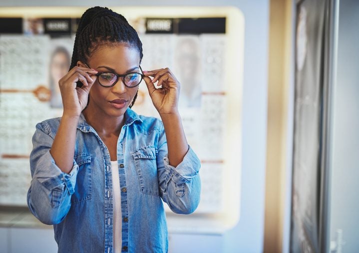 Single beautiful young African-American woman in blue shirt testing the fit for eyeglasses on her head in eye wear shop