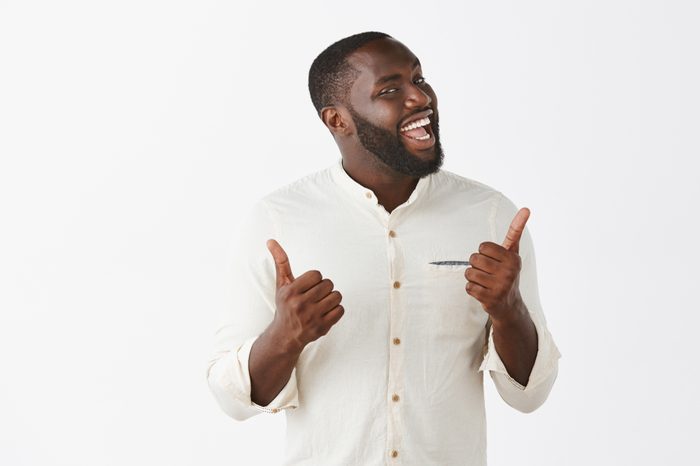 Indor shot of funny and emotive joyful dark-skinned guy with beard in trendy outfit, showing thumbs up and turning face at camera, smiling broadly, liking great and awesome party over gray background