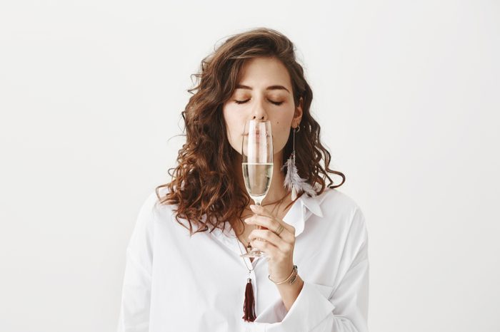 Studio portrait of beautiful sensual caucasian brunette smelling white wine while holding glass near nose, standing with closed eyes and enjoying taste over gray background.