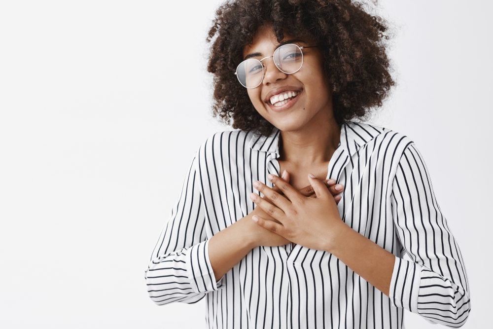 Close-up shot of touched and pleased happy dark-skinned girl with afro hairstyle in glasses and striped blouse holding hands on chest and smiling broadly being pleased receiving compliments
