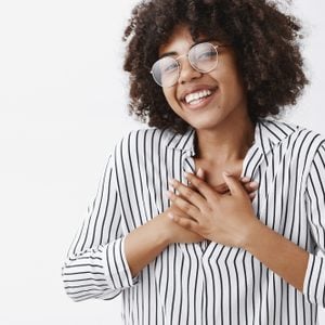 Close-up shot of touched and pleased happy dark-skinned girl with afro hairstyle in glasses and striped blouse holding hands on chest and smiling broadly being pleased receiving compliments