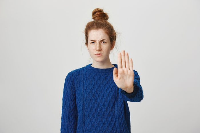 I am not interested live me alone. Portrait of annoyed angry good-looking redhead female in winter sweater pulling palm at camera with stop or no gesture, displeased while standing over gray wall