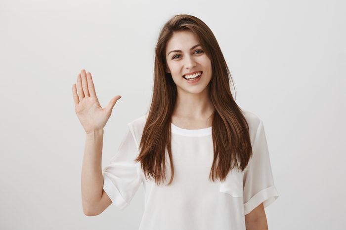 Friendly woman says hi to new neighbours. Portrait of charming young emotive woman waving with raised hand, greeting or welcoming close friend, smiling broadly at camera, standing over gray wall
