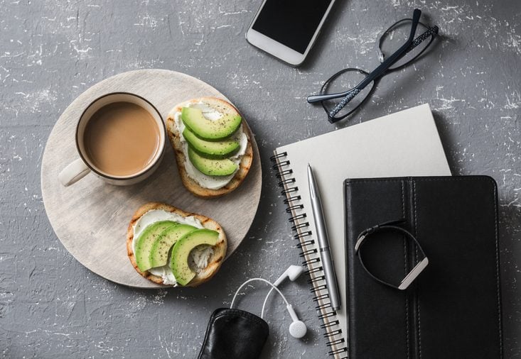 Coffee break during working hours. Flat lay business workplace with notebook, tablet, phone, glasses. Coffee and sandwich with cream cheese and avocado on a gray background, top view
