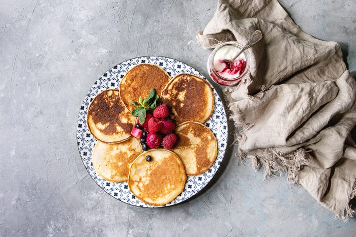 Homemade pancakes served on decorate plate with berries, mint, glass jar of yogurt and cloth over grey texture background. Top view, space.