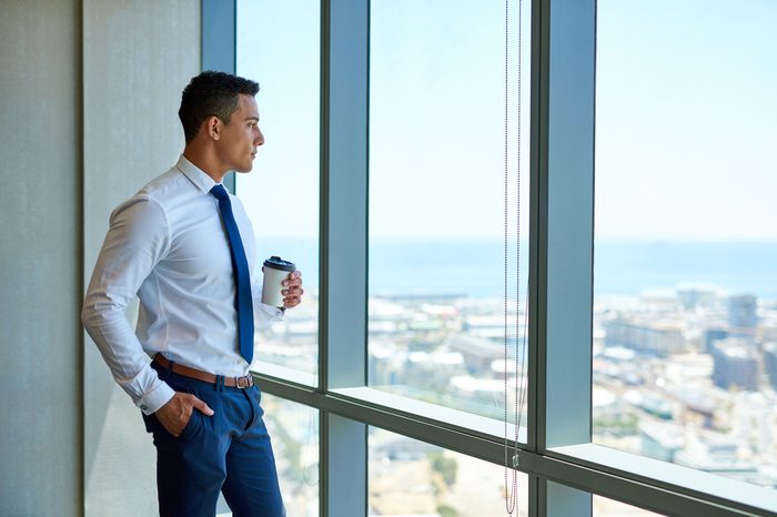 Confident young businessman drinking a cup of coffee while looking through windows at the city from high up in an office building