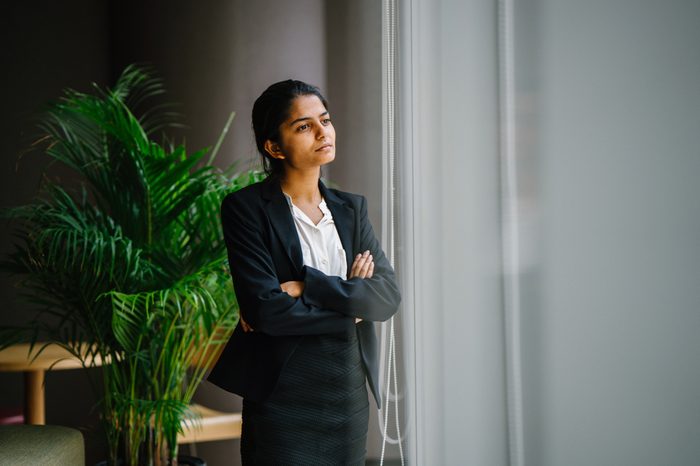 Portrait of a young Indian Asian business woman standing by the window in a meeting room, smiling with her arms crossed. She looks confident, happy and optimistic. 