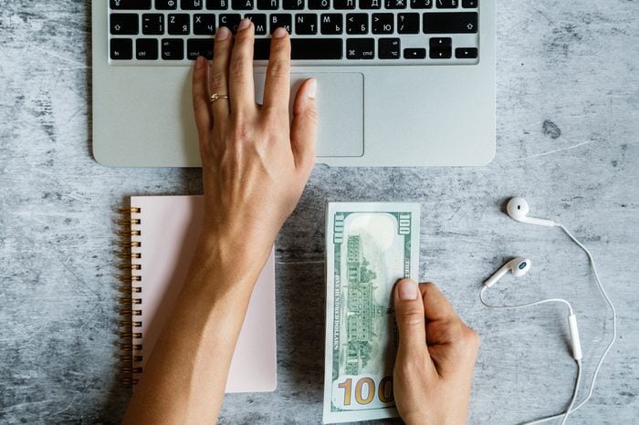 Desktop flatlay: laptop, notebook, woman’s hand holding cash money, left hand typing on keyboard, headphones lying on gray marble background