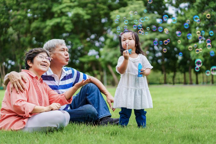 Asian cute granddaughter making bubbles and attractive happy grandparents look at her in the public garden at vacation day or holiday. Family Concept.