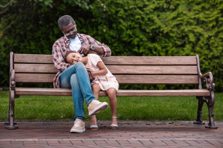 adorable african american girl and her grandfather sitting on bench in park 