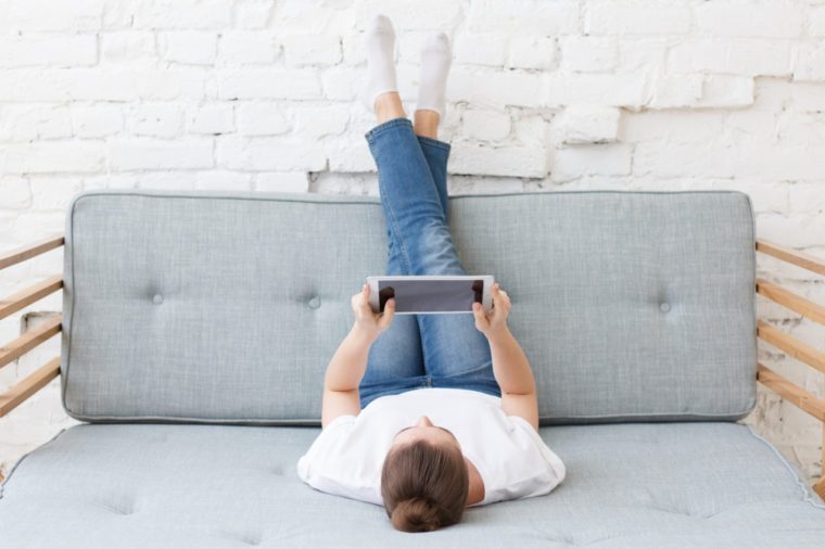 Young woman laying on couch in loft interior, watching movie or playing video game with her tablet pc