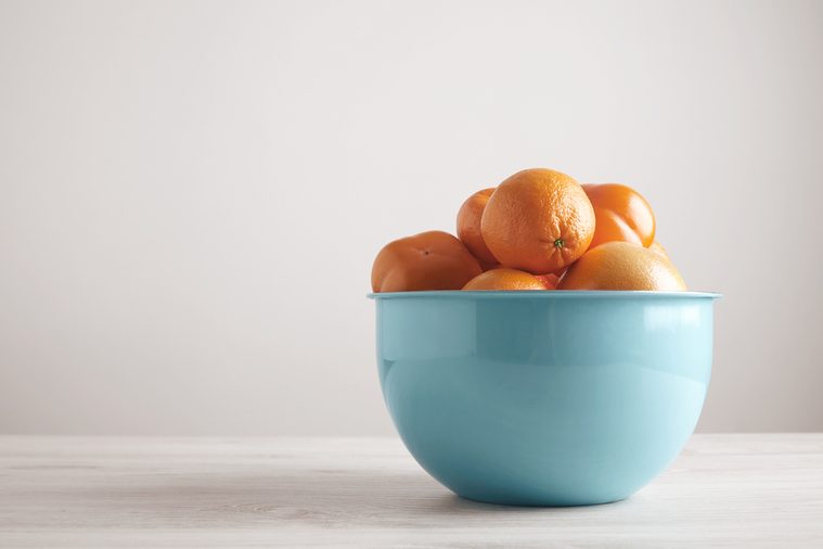 Different fruits and citrus in big metallic blue bowl in front of blank wall on white wooden table from side