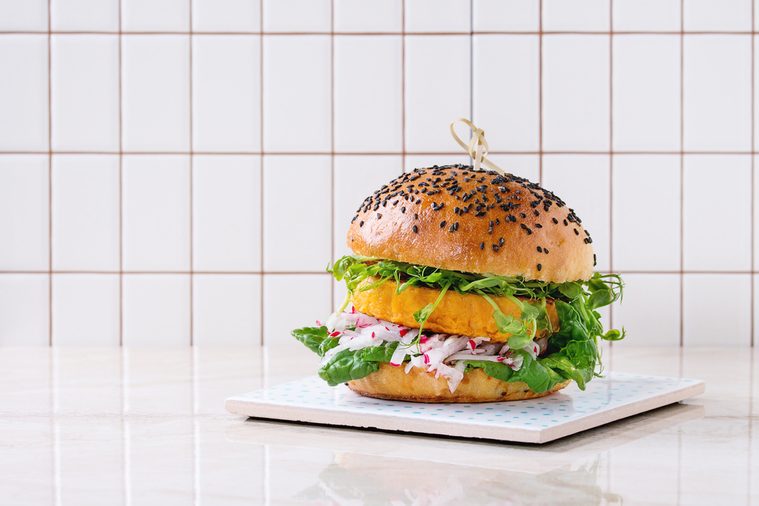 Homemade veggie sweet potato burger with fresh radish and pea sprouts served on ceramic board over white marble table with tile wall at background.