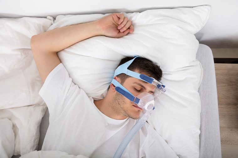High Angle View Of Man Lying On Bed With Sleeping Apnea And CPAP Machine