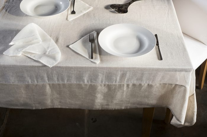 Shot of corner of dining table covered with white linen and with a crumpled table napkin and two sets of dinnerware arranged on opposite sides.