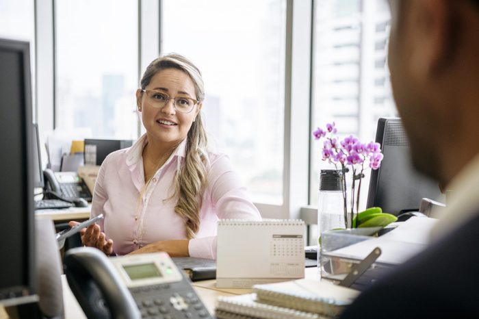 Office worker in her 30s siting at desk and looking towards co-worker, listening and smiling, two people meeting in office