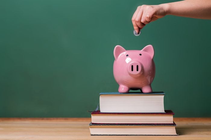 Person depositing money in a pink piggy bank on top of books with chalkboard in the background as concept image of the costs of education 