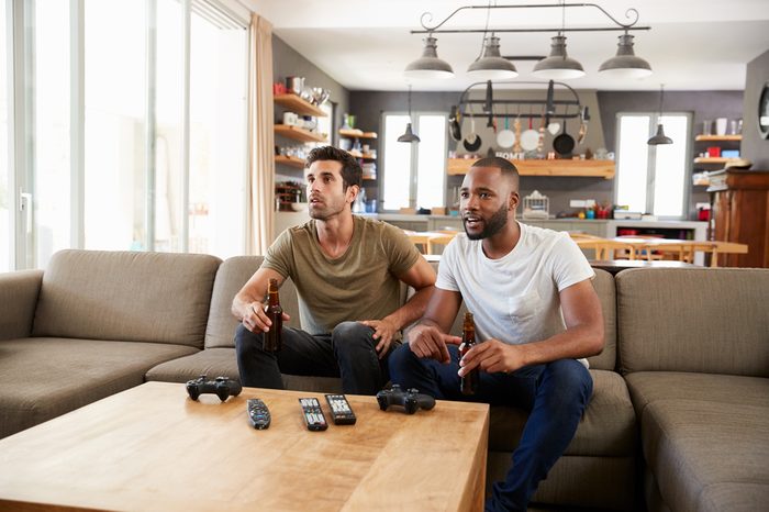 Two Male Friends Sit On Sofa And Watch Sports On Television
