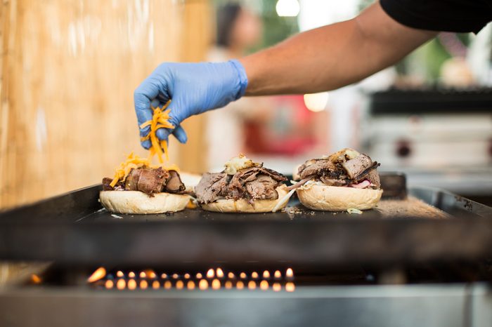 Crop faceless shot of person in gloves pouring cheese on burgers.