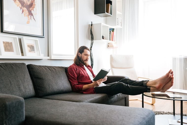 Handsome bearded young man sitting and reading book on sofa at home
