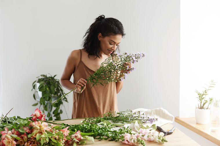 Attractive african female florist smiling making bouquet at workplace over white wall.