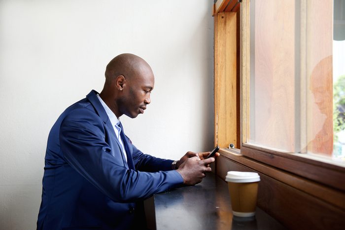 Side portrait of black businessman sitting in a cafe sending text message on cell phone 