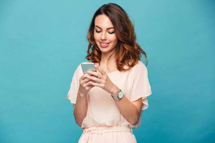 Contented smiling woman typing text message or scrolling through social networks using smartphone isolated over blue background