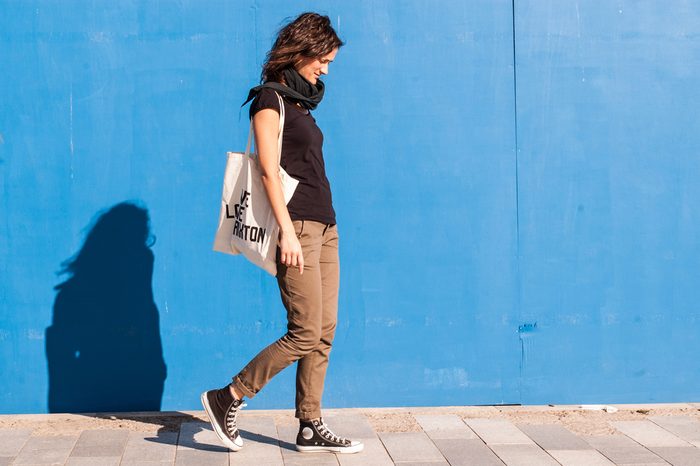 Young girl dressed in casual brown chinos, black sneakers and T-shirt walking on a street with blue wall in background.