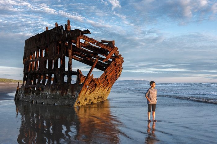 Boy stands by the Peter Iredale shipwreck in the golden sunset light in northwest Oregon.