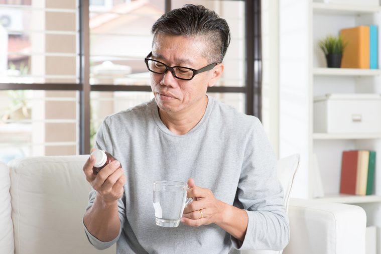 Men health concept. Portrait of 50s mature Asian man reading the label on bottle medicine, sitting on sofa at home.
