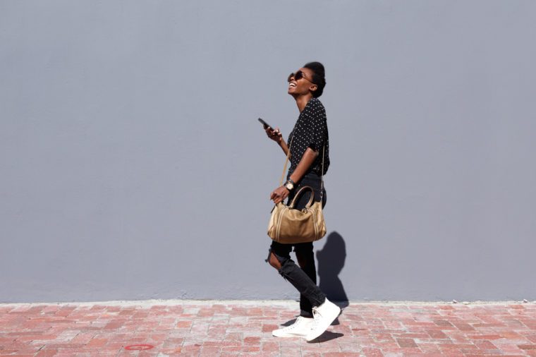 Full length side view portrait of smiling young black woman walking outdoors with mobile phone