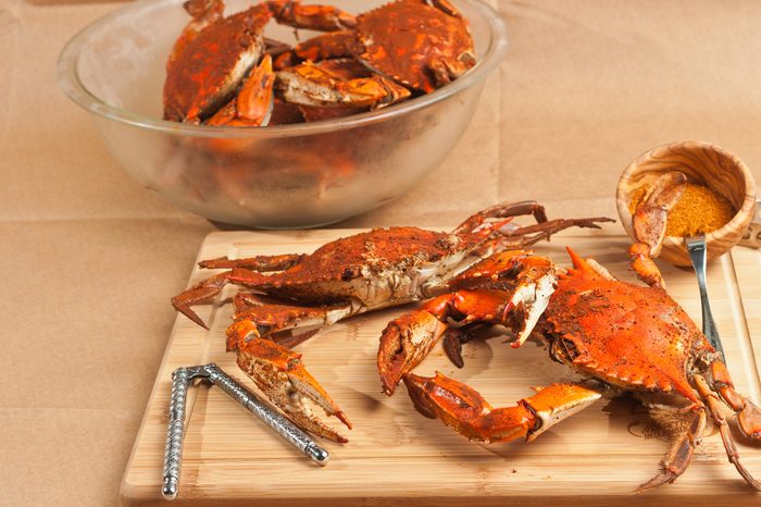  Two steamed, seasoned, male jumbo Chesapeake Bay blue claw crabs on a wood cutting board with nut cracker and a wood bowl of seasoning and a clear glass bowl of crabs on brown paper table cloth