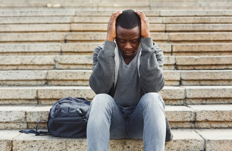 Desperate african-american student sitting on stairs outdoors in university campus. Man raised hands to his head. Migraine, education and overworking concept, copy space