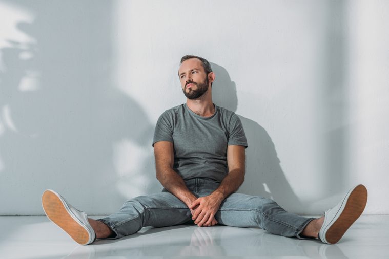 full length view of depressive bearded middle aged man sitting on floor and looking away 