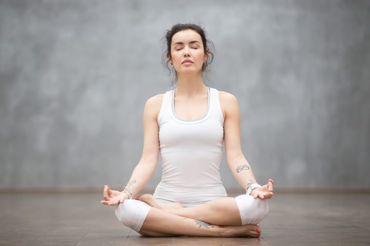 Front view portrait of beautiful young woman with floral tattoos working out against grey wall, resting after doing yoga exercises, sitting in ardha Padmasana, Lotus pose, relaxing. Full length